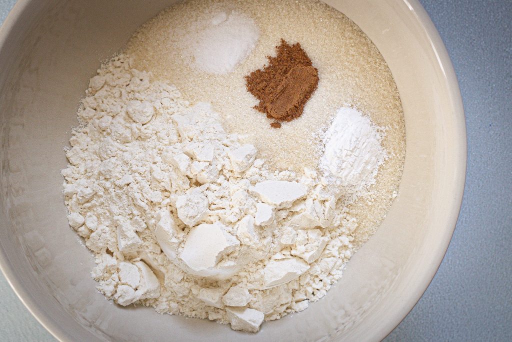 Dry ingredients for how to make strawberry muffins recipe. White ceramic bowl filled with all-purpose flour, raw sugar, Ceylon cinnamon, aluminum free baking powder, and sea salt