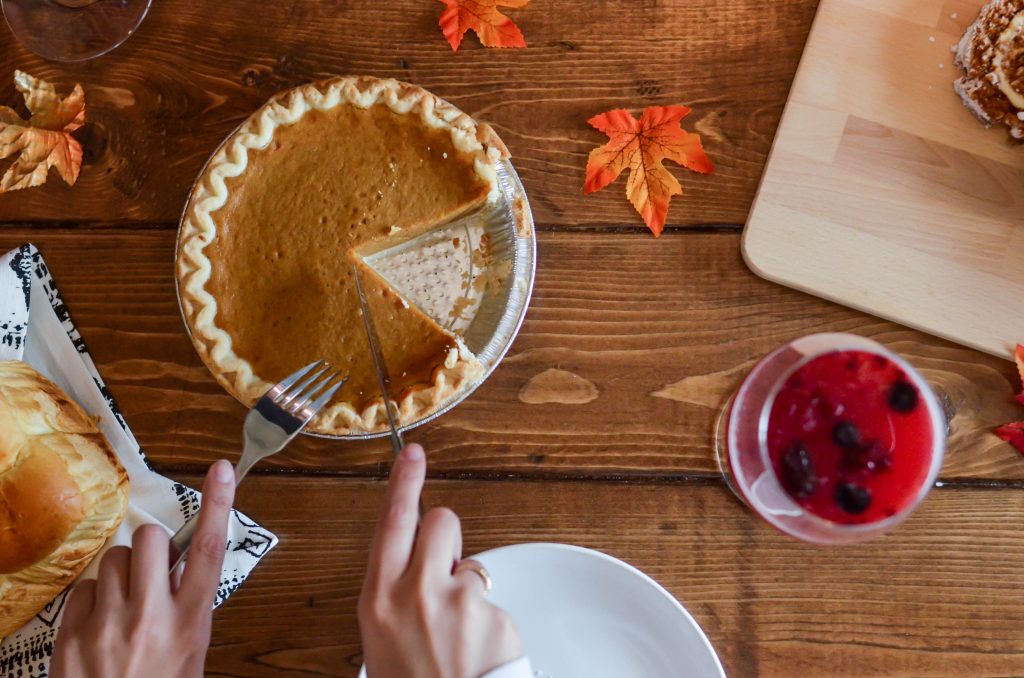 hands cutting a pumpkin pie surrounded by decorative orange leaves. a cranberry beverage is served beside it on the wooden table