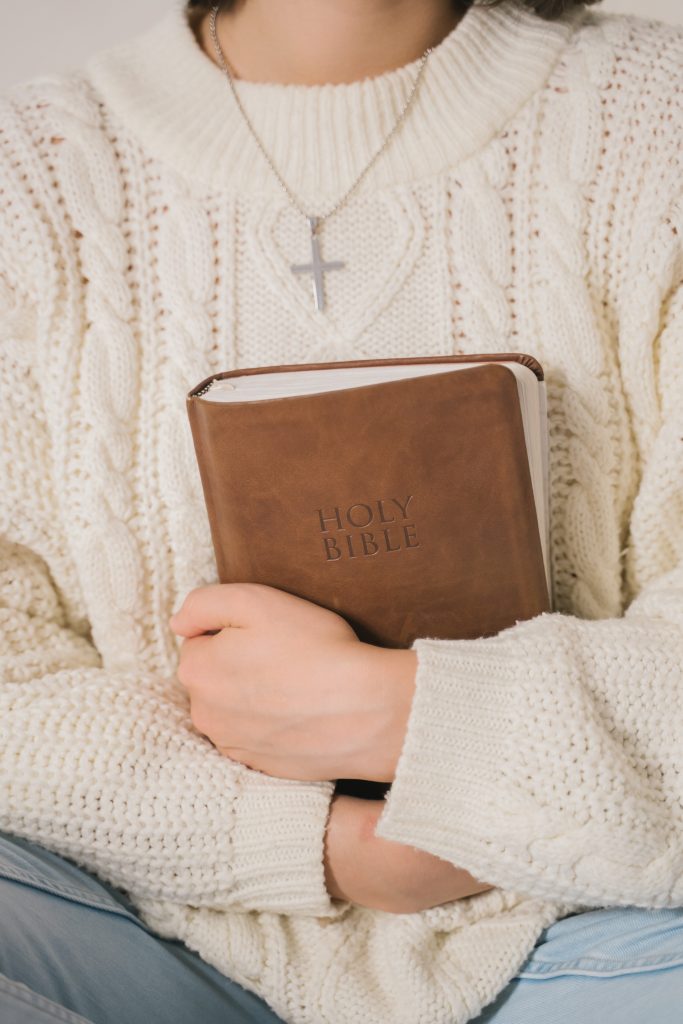 A woman wearing a cream chunky knit sweater adorned with a silver cross necklace. She holds a leather bound Holy Bible in her arms.